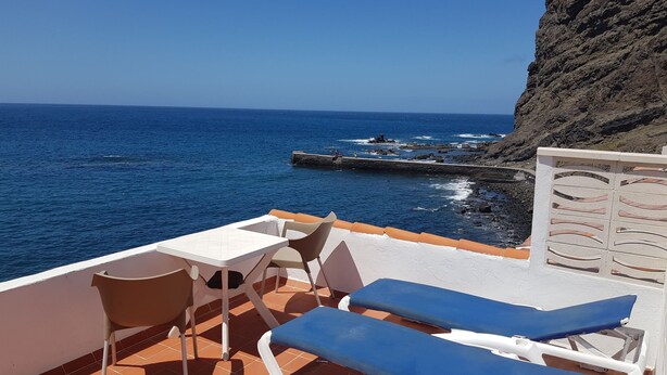 Een terras met een tafel en stoelen, uitkijkend op een rotsachtige kustlijn en de zee.