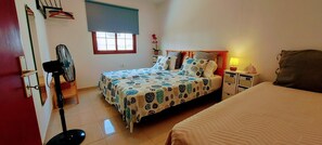 Ein Schlafzimmer mit Bett, Nachttisch mit Lampe, Standventilator, Fenster mit Jalousie und einer Zimmerpflanze.