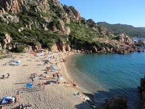 A beach with people sunbathing and swimming, surrounded by rocky cliffs and greenery. - Villa Ginepro (Valledoria)
