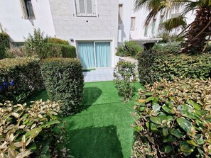 A courtyard with artificial green turf, trimmed hedges, and a building with white walls and shuttered windows.
