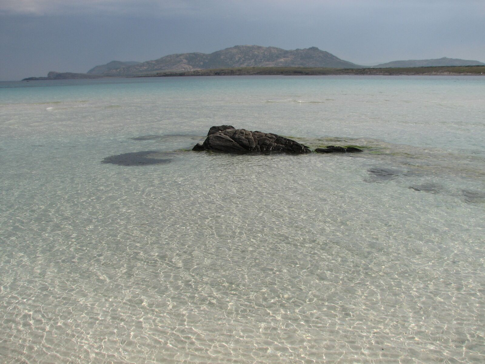 A clear shallow water body with a rock formation in the center and distant mountains.