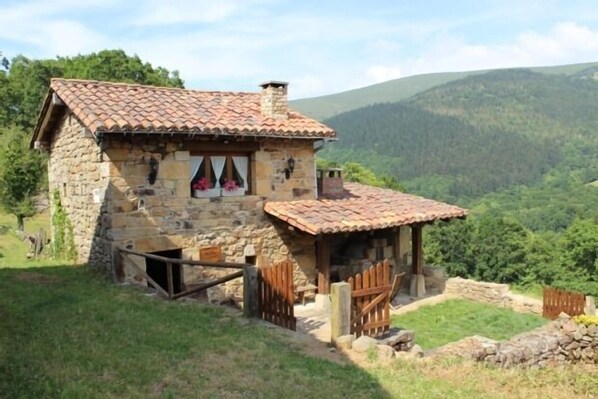 A stone house with a tiled roof, surrounded by a stone wall and wooden gates, set against a mountainous backdrop.