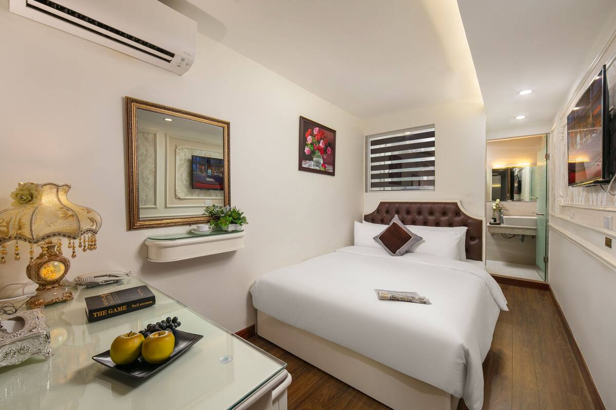 Una camera d'albergo con un letto, comodino, lampada, specchio e un piccolo ripiano con oggetti decorativi.