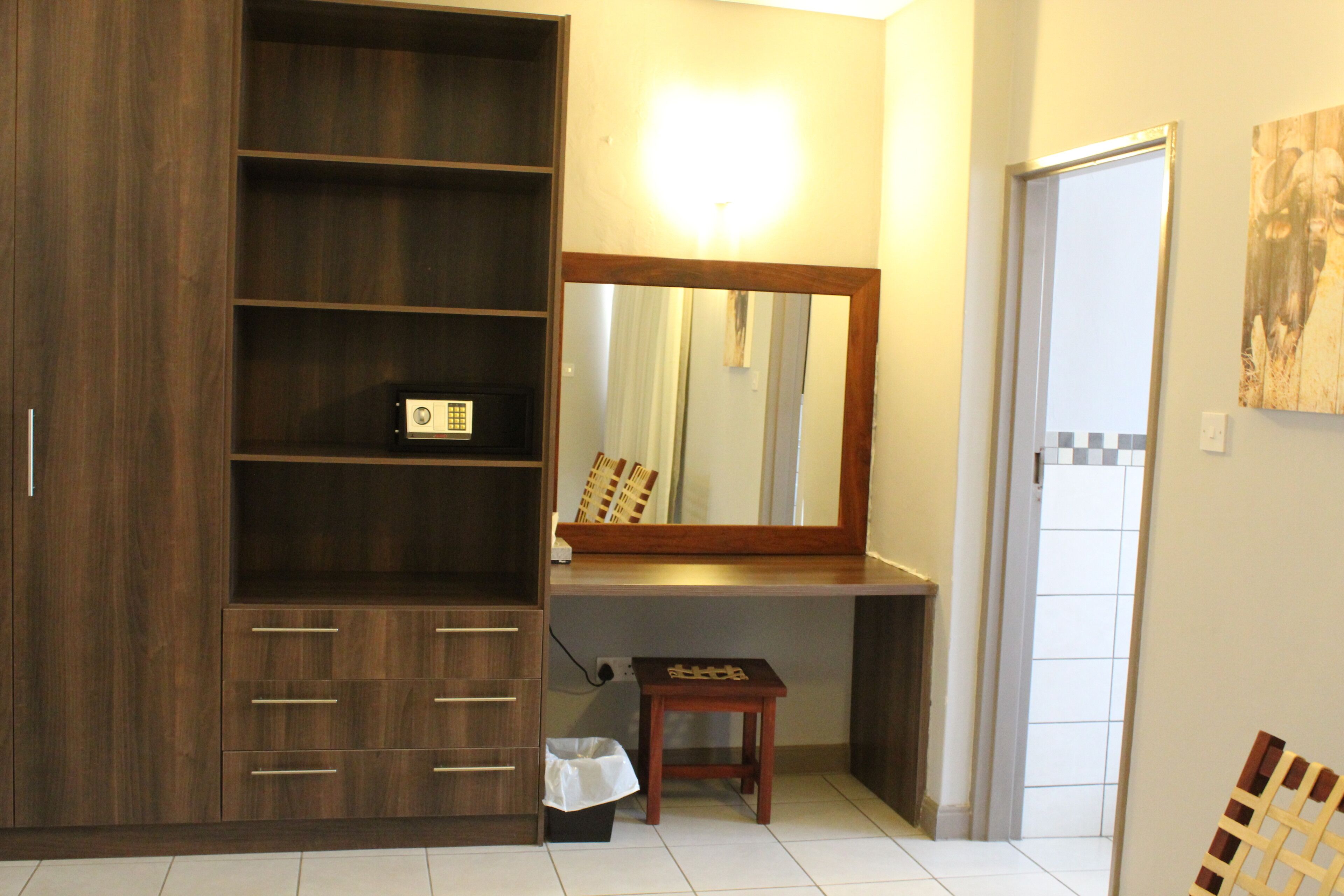 A room with a wooden wardrobe, a mirror, a small table, and a trash bin.