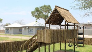 A wooden play structure with a ramp and a platform, surrounded by a fence and a grassy area. - Tlou Safari Lodge (Kasane)