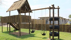A wooden play structure with swings and a slide, surrounded by a fence and houses in the background. - Tlou Safari Lodge (Kasane)