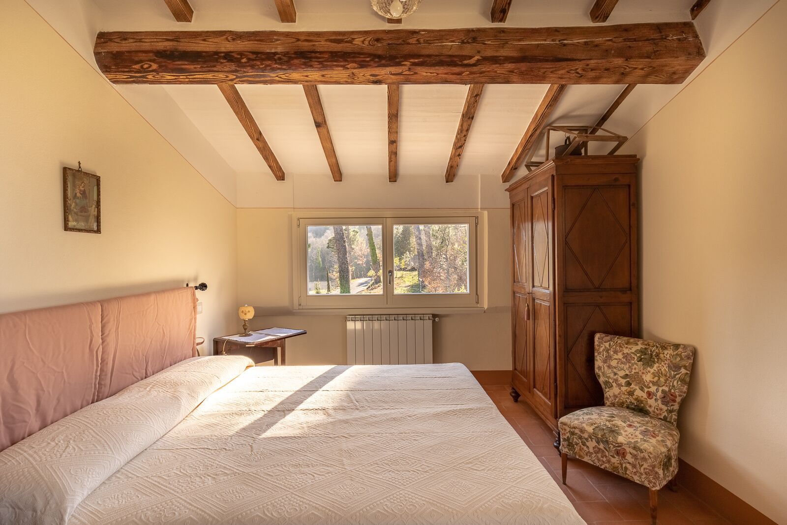 Une chambre avec un grand lit, une armoire en bois, une chaise, une fenêtre donnant sur des arbres et un radiateur.