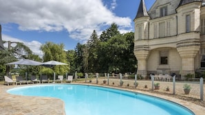 Una piscina con sillones de descanso y sombrillas, rodeada por una terraza de piedra y un edificio grande con una torre.