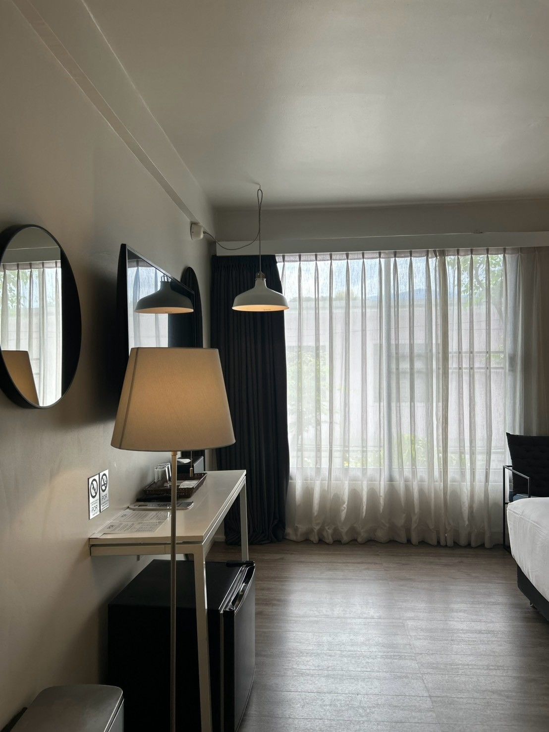 A hotel room with a bed, a desk with a lamp, a television, and a large window with curtains.