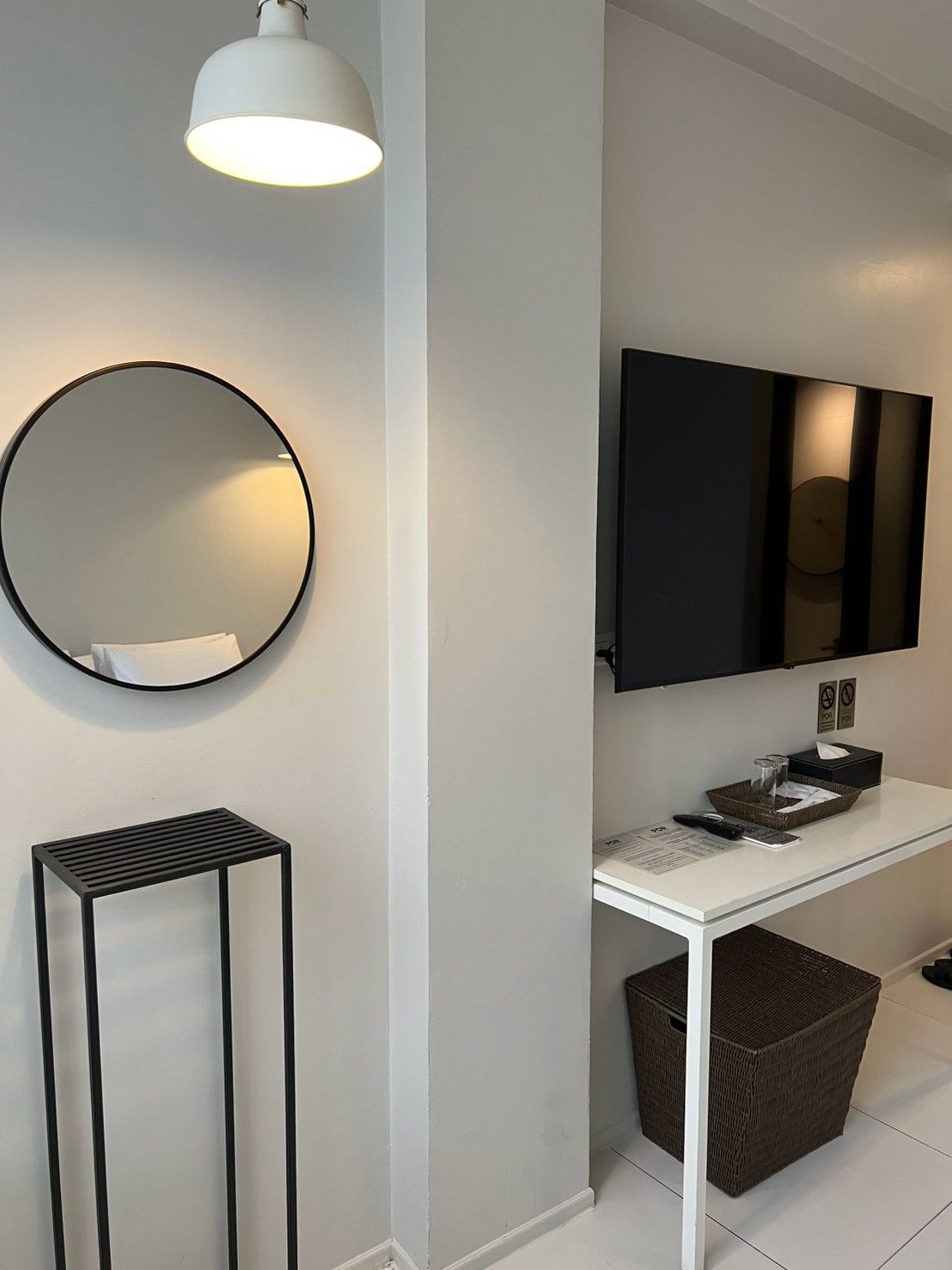 A hotel room with a white console table, a round mirror, a black-framed TV, and a black floor lamp.