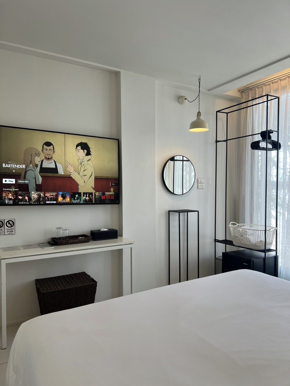 A hotel room with a large TV displaying a menu, a bed, a bedside table, a small desk with a basket, a round mirror, and a hanging light fixture.