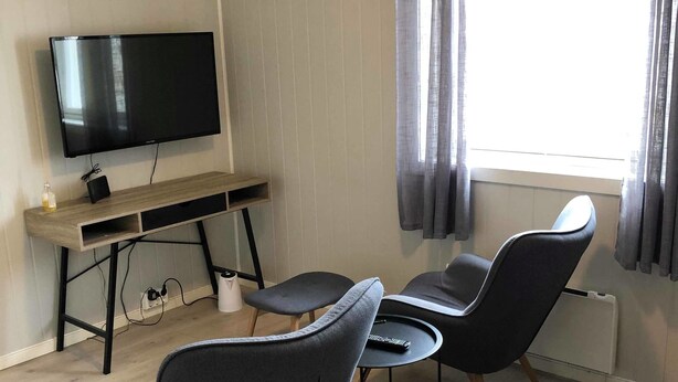 A modern room with a flat-screen TV mounted on the wall, a wooden desk with a chair, and two grey armchairs around a small round table. - Mosjøen Hybelhus (Vefsn)