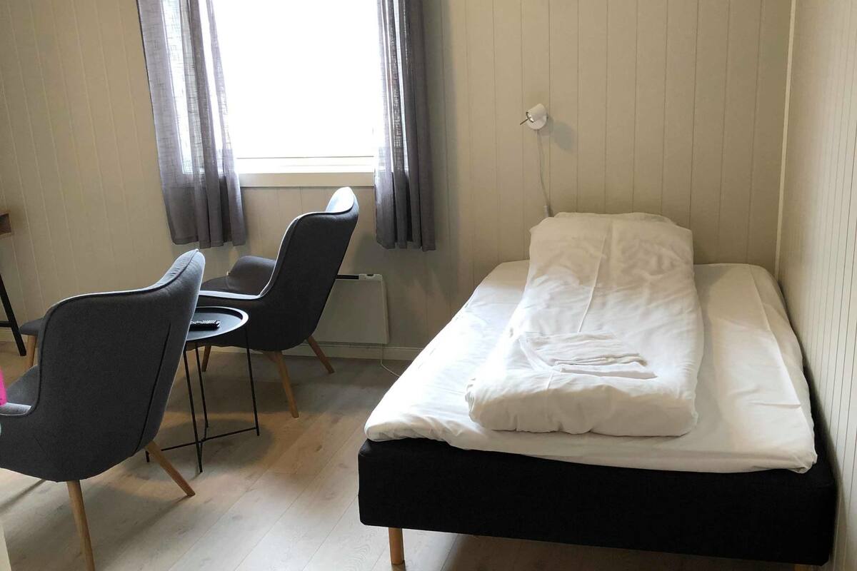 Une petite pièce, bien rangée, avec un lit, deux chaises, une petite table et une fenêtre avec des rideaux.