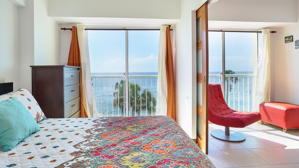 Un dormitorio con cama, cómoda, una silla roja y un balcón con vista al mar.