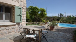 En stenbelagt terrasse med et rundt bord og stole, en stenvæg, en pool og frodig beplantning omkring.