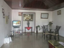 Una sala de estar con televisión, cuadros enmarcados, una mesa de comedor con sillas y una mesita con un florero.