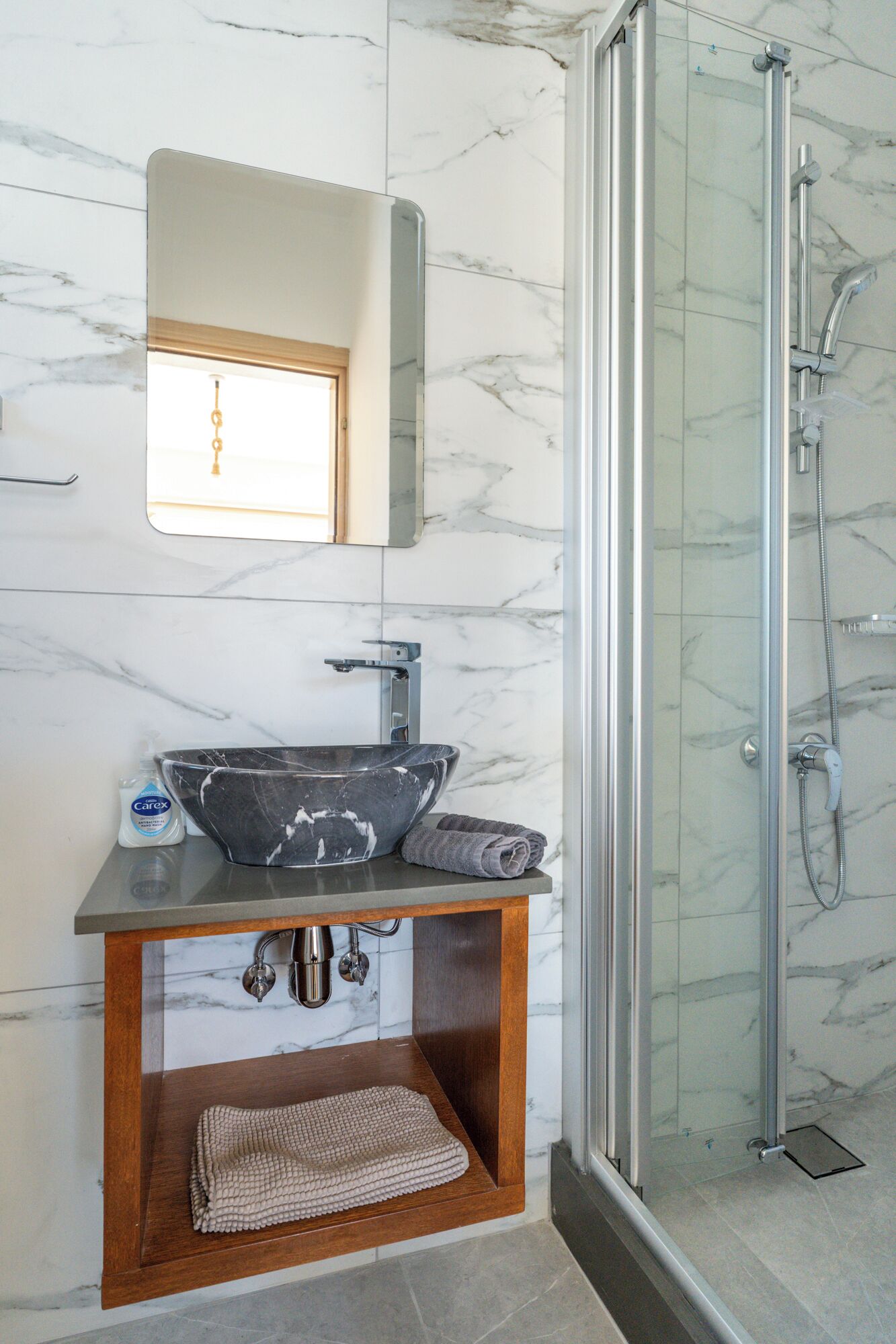 A bathroom with a marble sink, a wooden vanity, a glass shower enclosure, and a marble-tiled wall.