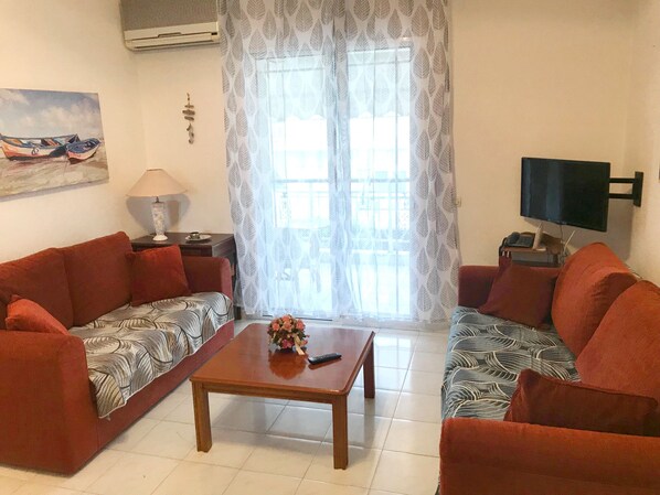 Ein Wohnzimmer mit zwei roten Sofas, einem Holztisch, einem Fernseher auf einem Standfuß und einem Fenster mit gemusterten Vorhängen.