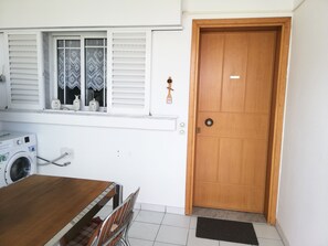 Ein Zimmer mit Holztür, einem Fenster mit gemusterten Vorhängen, einer Waschmaschine und einem Esstisch mit Stühlen.