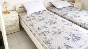 A room with two beds, each with a floral-patterned mattress and white pillows. There are bedside tables with lamps on either side of the beds. - Hanioti Apartment (Kassandra)