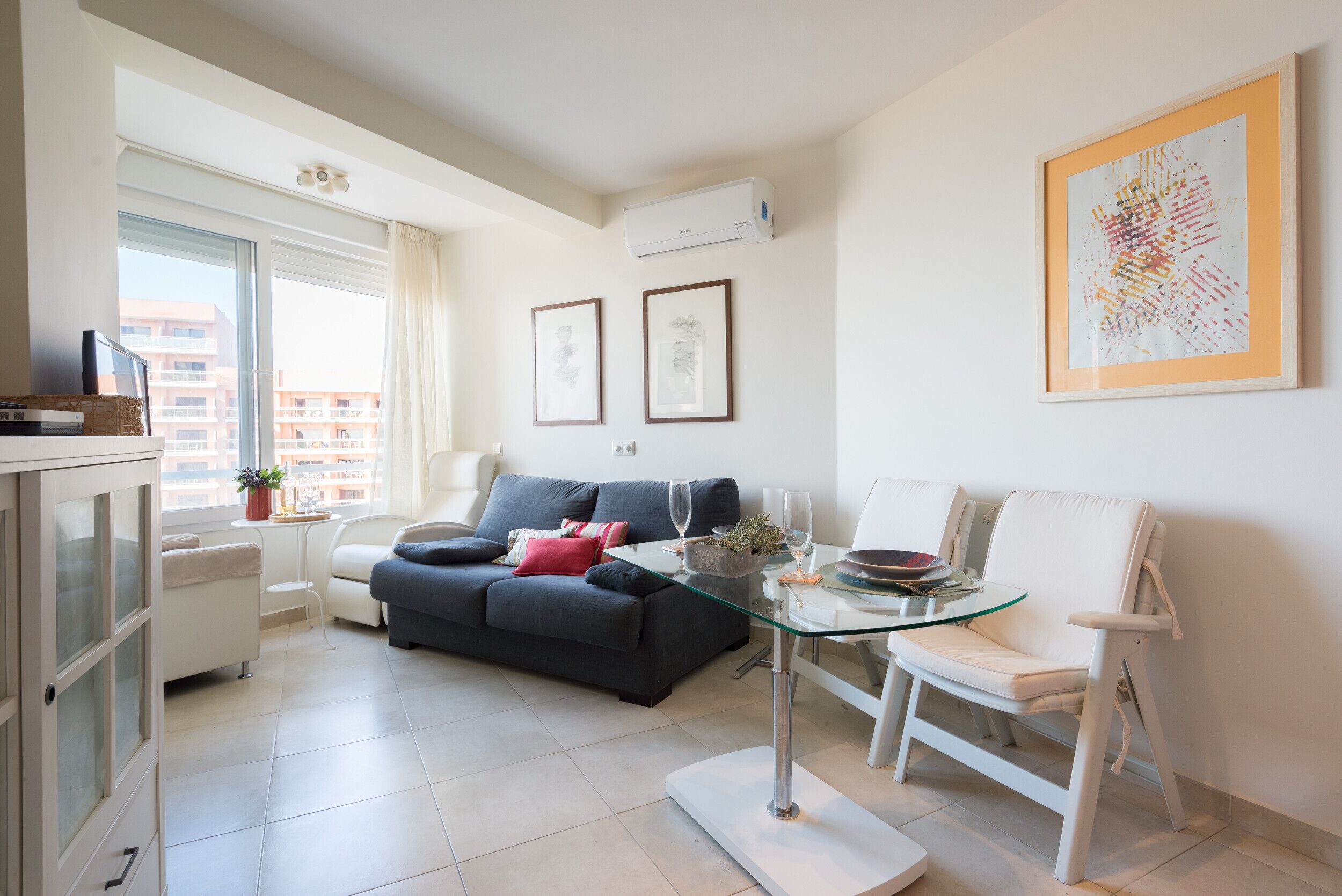 A modern living room with a glass coffee table, a dark sofa, a white armchair, and a small side table with a plant.
