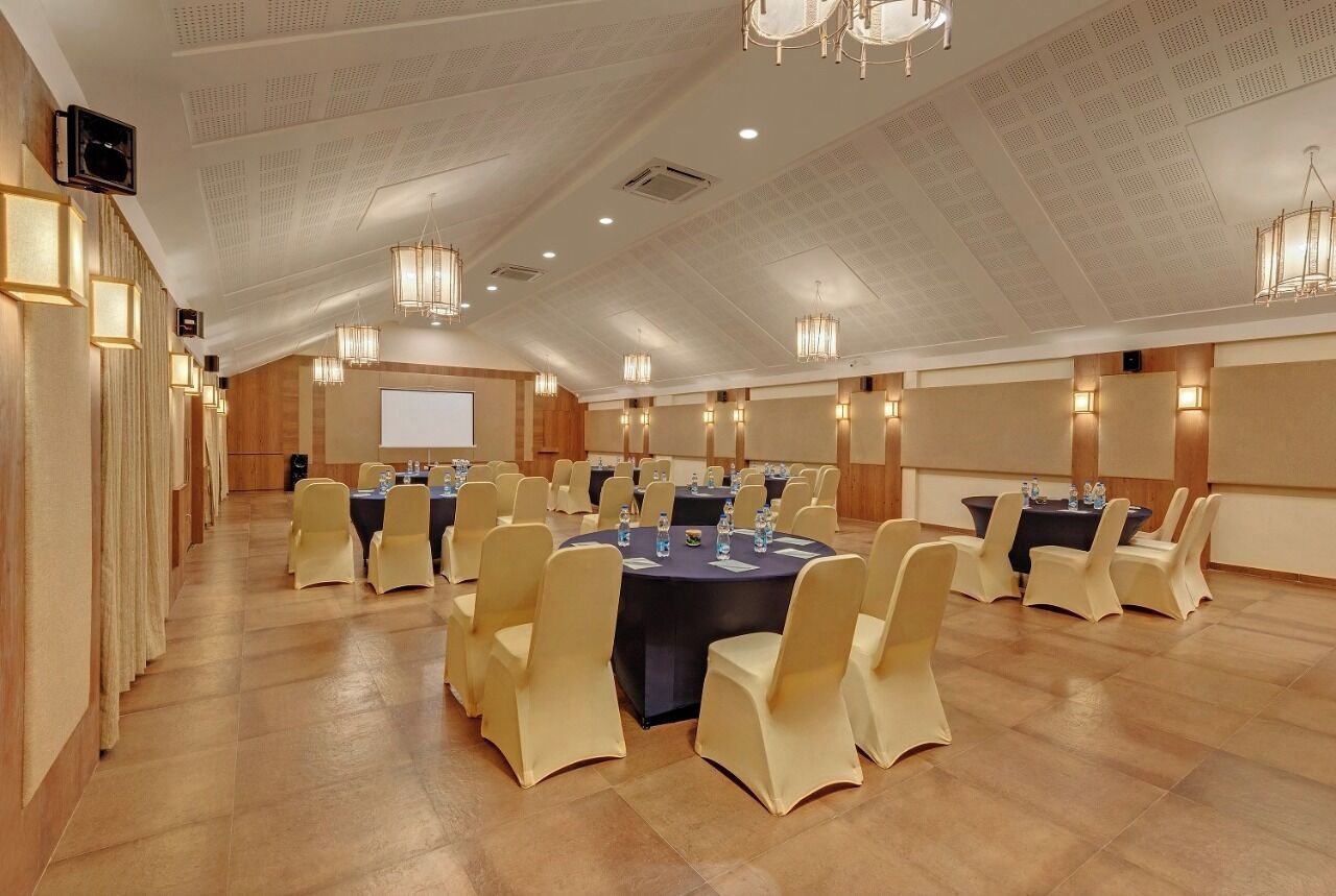 Un salón de conferencias con mesas redondas, sillas color beige y una pantalla de proyector.