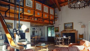 Ein großzügiges Wohnzimmer mit hohen Decken, Holzbalken und einem Kamin. Es gibt mehrere Sitzbereiche und eine Treppe, die in einen oberen Bereich führt.