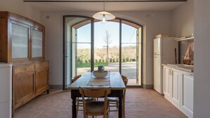 Een keuken met een eettafel, stoelen, een hanglamp en uitzicht door een grote glazen deur.