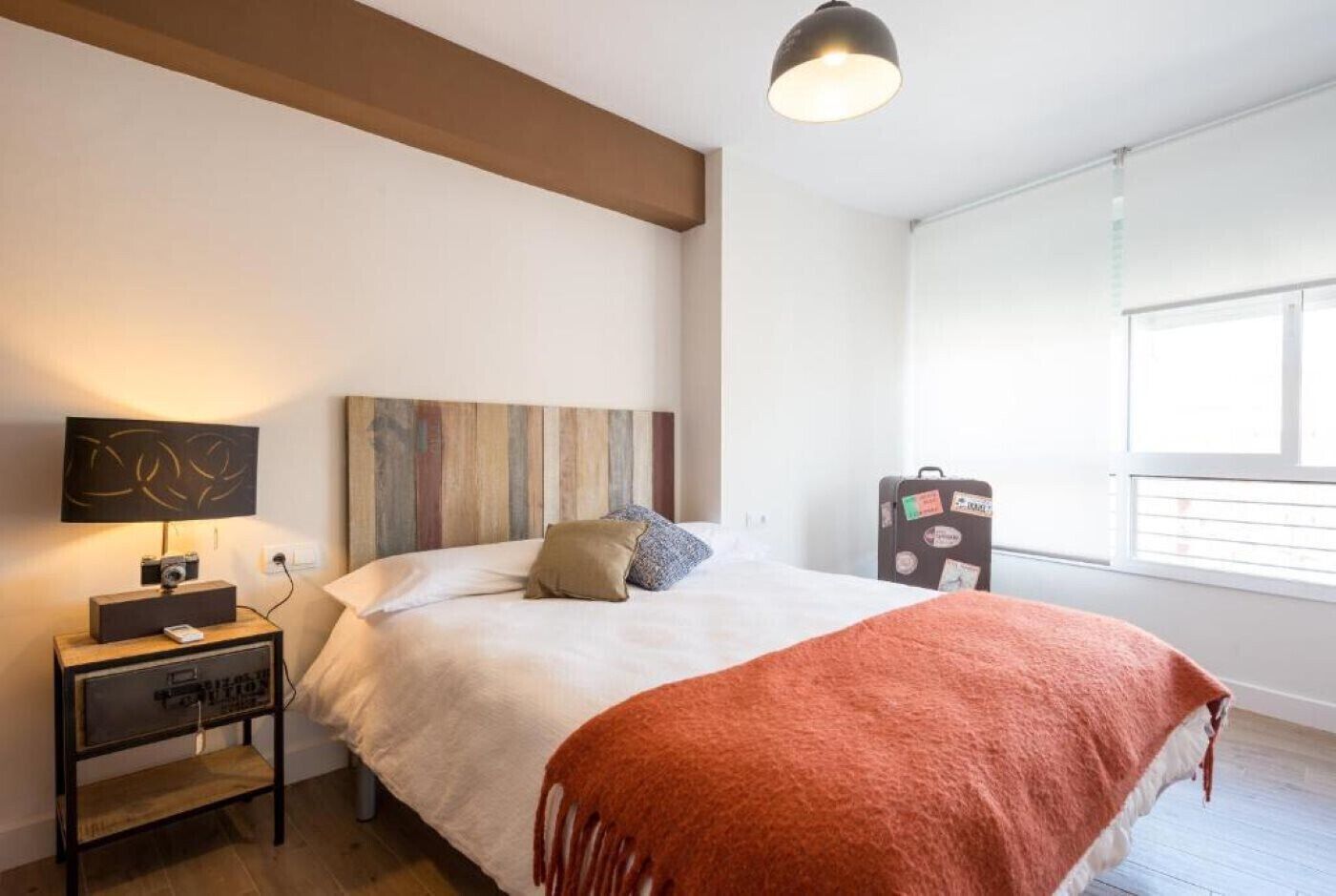 A bedroom with a bed, a nightstand, a lamp, a suitcase, and a window.