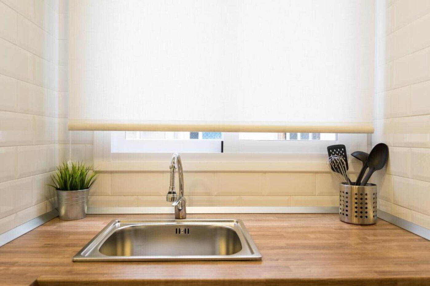 A kitchen with a stainless steel sink, a window with blinds, a potted plant, and a utensil holder.