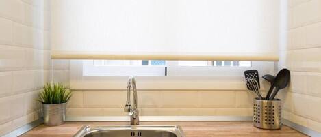 A kitchen with a stainless steel sink, a window with blinds, a potted plant, and a utensil holder.