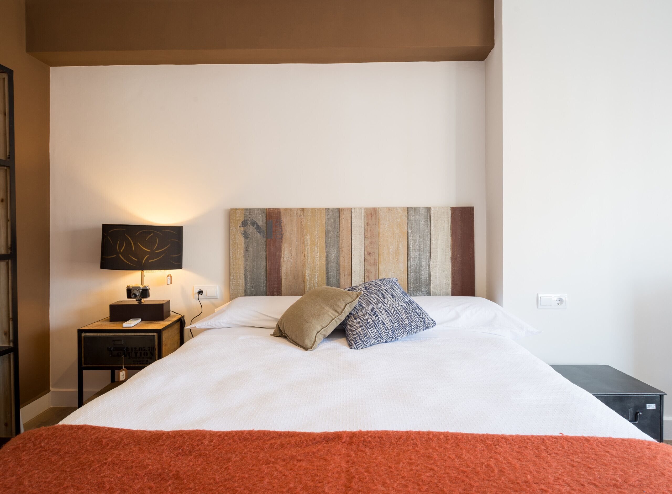 Una cama bien hecha con una manta blanca y naranja, un cabecero de madera, una lámpara de noche y una mesita con una cámara y un control remoto.