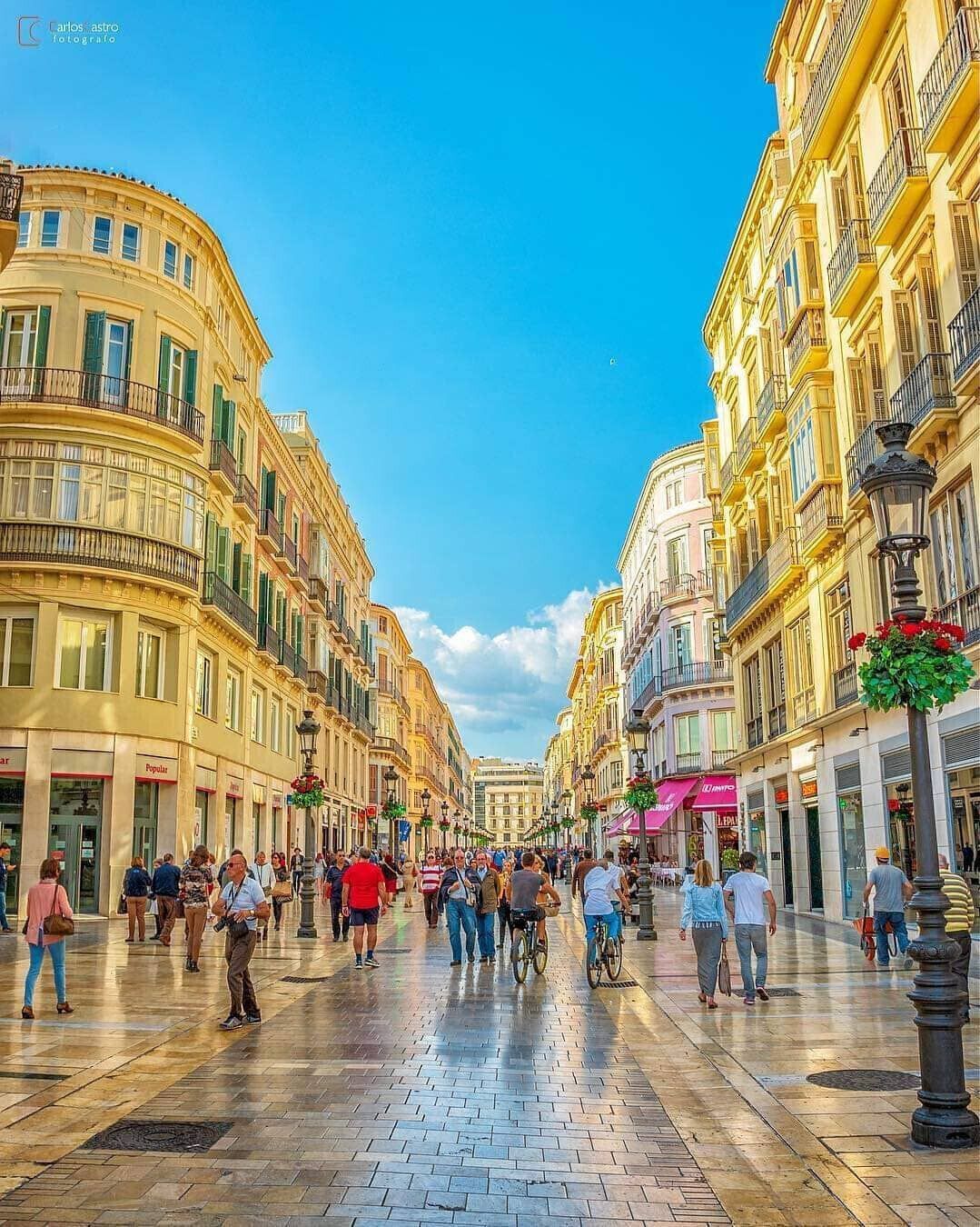 A wide pedestrian street lined with historic buildings, featuring a central open space with people walking and cycling.