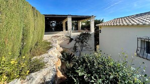 Uma área externa coberta com pérgola, um caminho de pedra e um edifício branco com telhado de azulejos.
