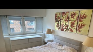 A bedroom with a bed, a window with blinds, a bedside table with a lamp, and a wall with three paintings. - Stunning self contained 1 bedroom flat in the heart of Putney.  (London)