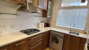 A compact kitchen with wooden cabinets, a gas stove, a built-in washing machine, and a window with blinds. - Stunning self contained 1 bedroom flat in the heart of Putney.  (London)