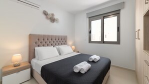 A modern bedroom with a tufted headboard, a large bed with a black quilted bedspread, two white pillows, and two rolled towels on the bed. There is a bedside table with a lamp, a wall-mounted air conditioner, and a window with a view of the outside. - Rhodes Luxury Apartments (Rhodes)