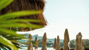 Blick auf einen Strand mit Liegestühlen und Sonnenschirmen, ein Pool und Berge im Hintergrund.