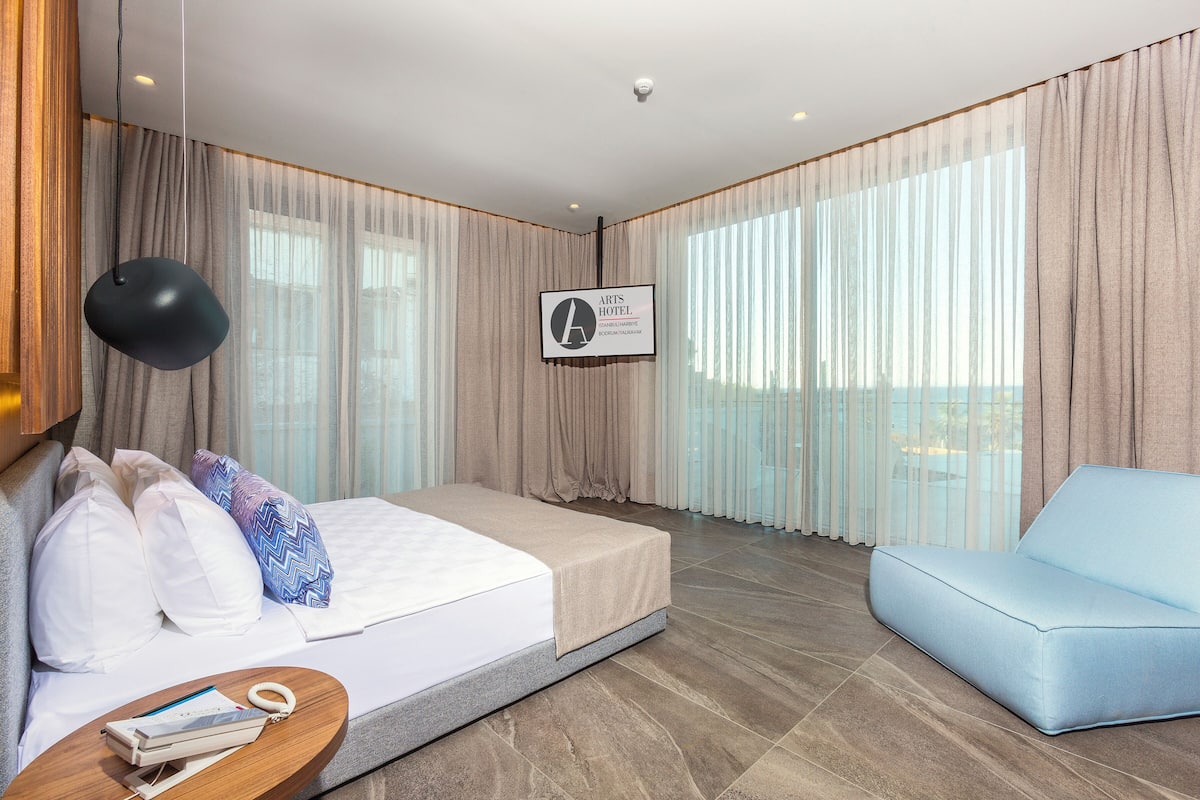 Habitació d'hotel moderna amb un llit, un sofà blau, una taula auxiliar de fusta i una finestra gran amb cortines.