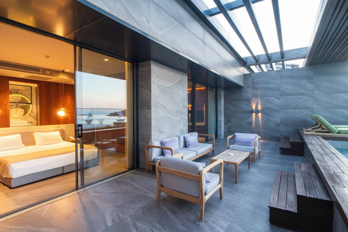 Habitació d'hotel moderna amb paret de vidre, un sofà, una banca de fusta i una taula petita.