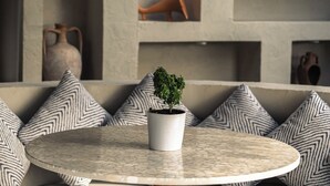 Ein runder Tisch mit einem Blumentopf, umgeben von gemusterten Kissen, vor einer strukturierten Wand mit Einbauten und einem Reetdach.