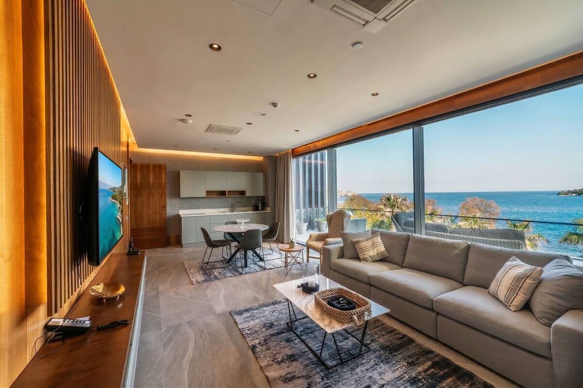 Un saló modern amb un televisor gran, un sofà, zona de menjador i vista al mar.