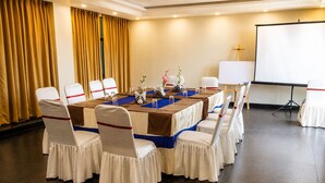 Une salle de conférence avec une longue table, des chaises recouvertes de housses blanches et ornées de rubans rouges, un écran de projection et une croix accrochée au mur.
