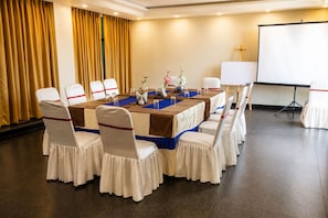 Ein Konferenzraum mit langem Tisch, Stühlen mit weißen Hussen und roten Bändern, einer Projektionsleinwand und einem Kreuz an der Wand.