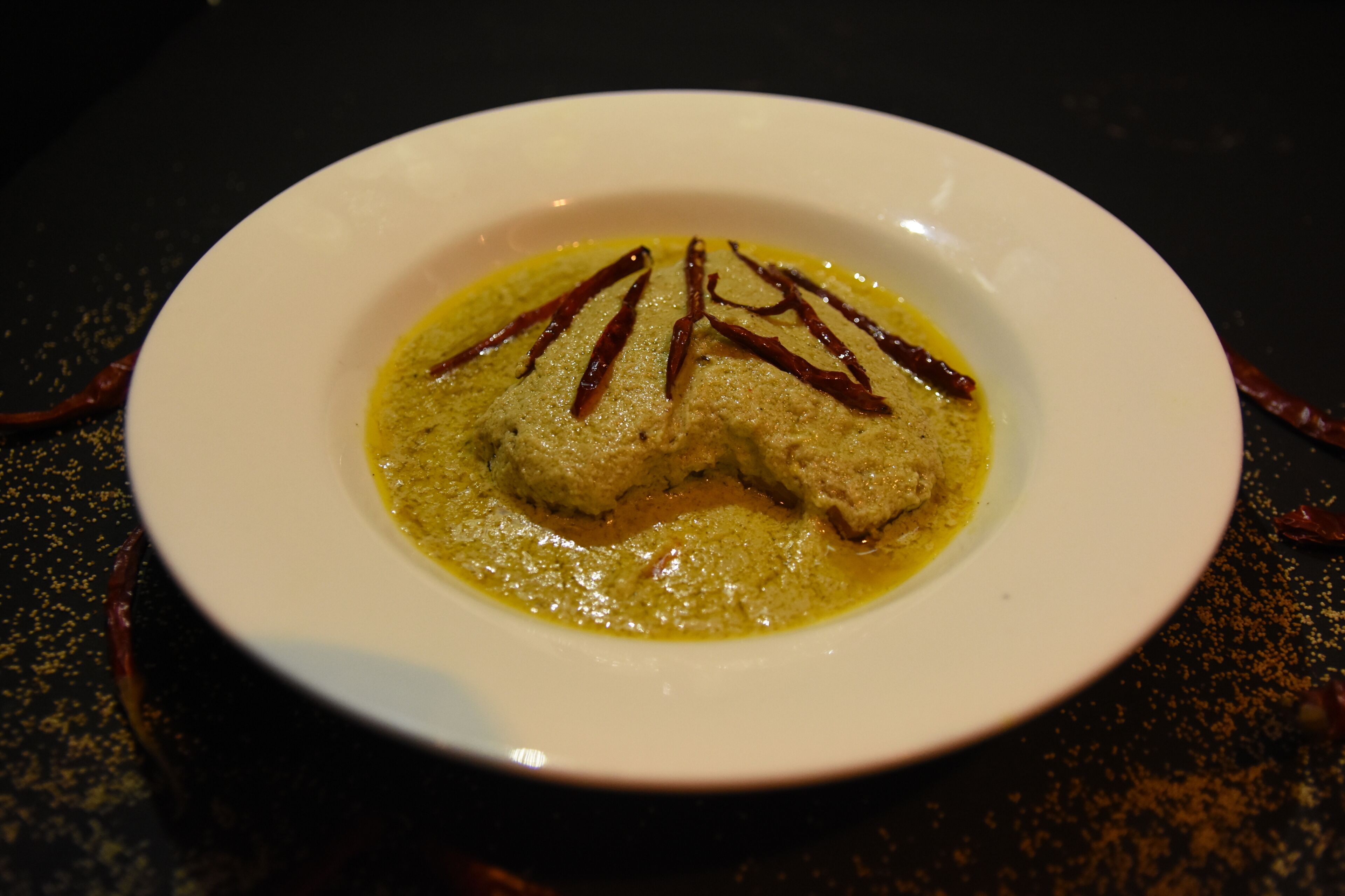 A white plate with a green curry and two pieces of meat, possibly chicken, garnished with red chili strips.