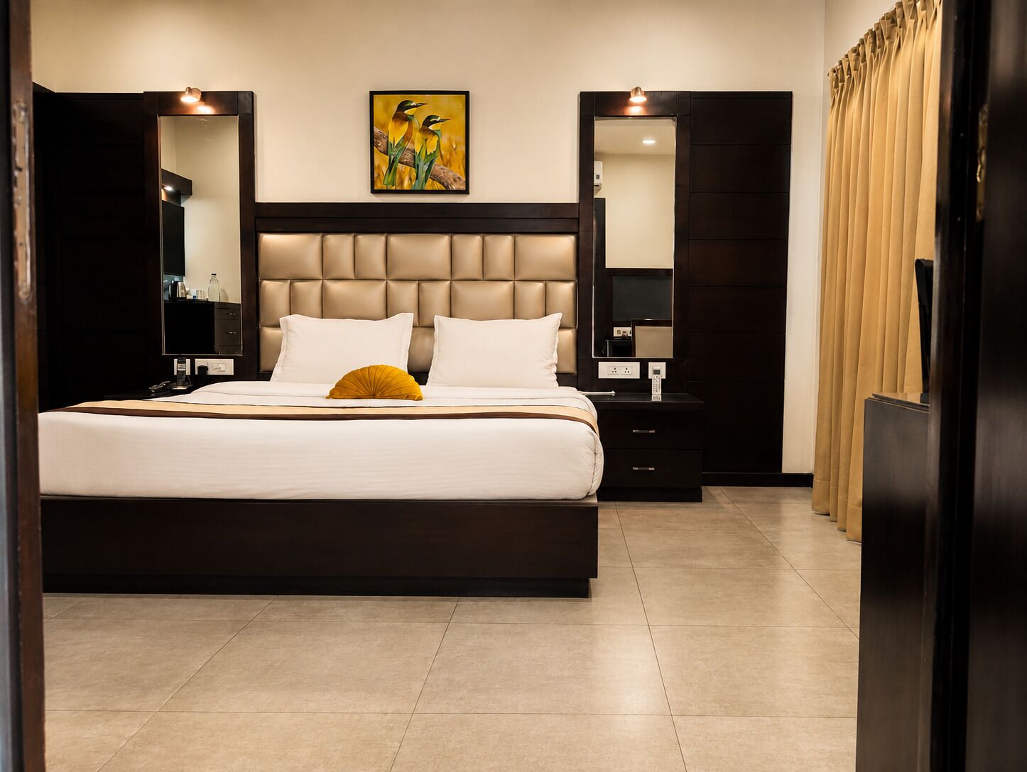 A modern hotel room with a large bed, a headboard, bedside tables, a mirror, and a painting on the wall.