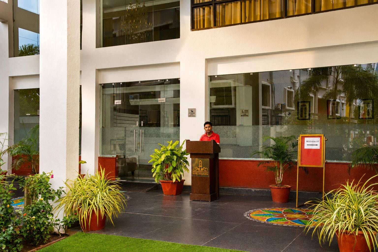 Vestíbulo de hotel con mostrador de recepción, plantas en macetas y un letrero que indica un restaurante.