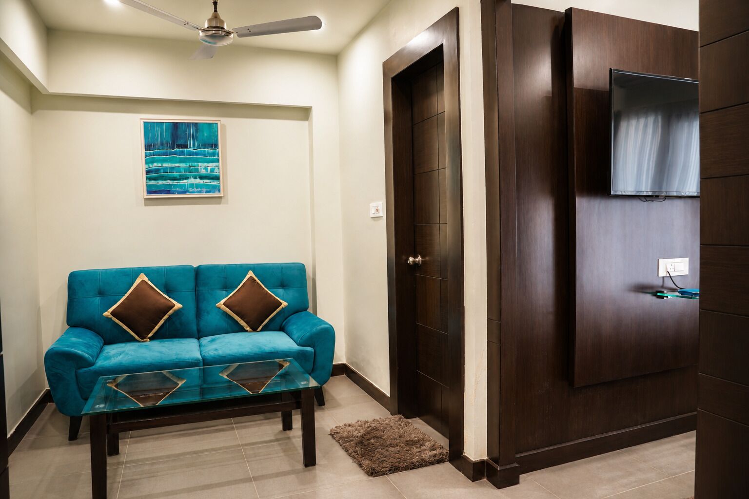 A room with a teal sofa, a glass coffee table, a wooden door, and a ceiling fan.