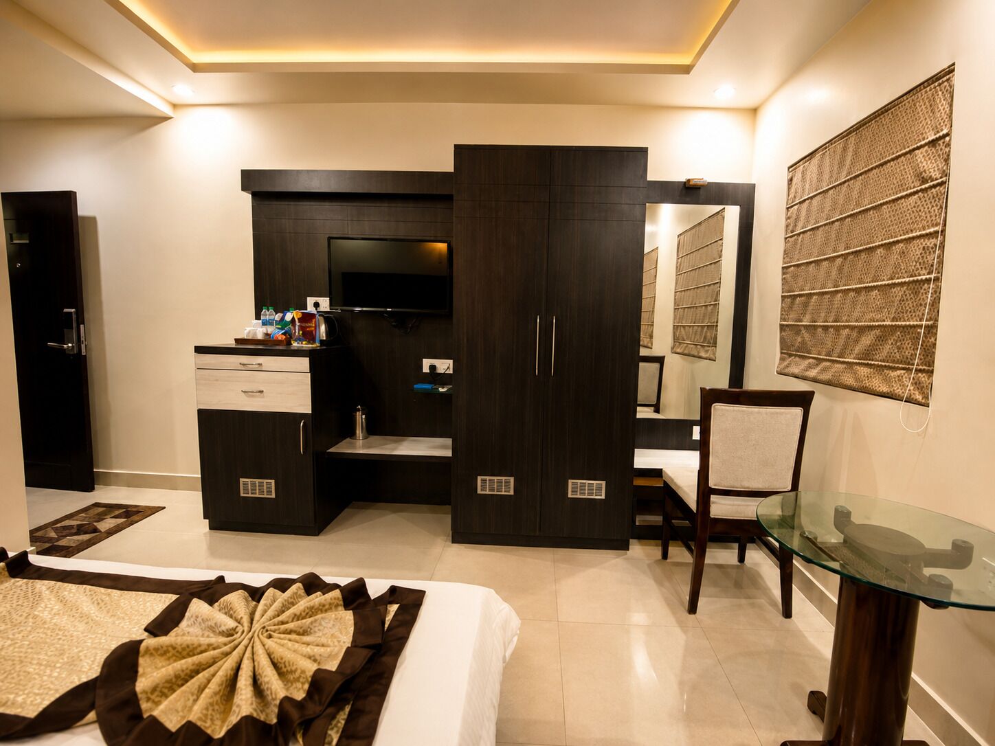 A hotel room with a bed, a desk, a wardrobe, a television, and a glass table.