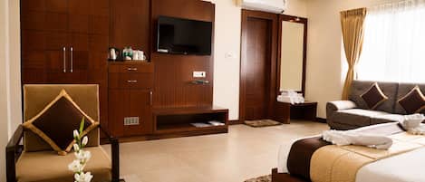 Habitación de hotel con cama, sofá, televisor, armario y una mesita con un florero.